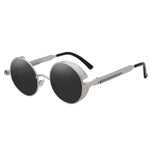 Stylish sunglasses with a black lens and a silver rounded frame. The edges of the frames are extended by a small stylish bezel to cover the top and bottom of the eyes. With a spring attached across to the lower part of the handles.